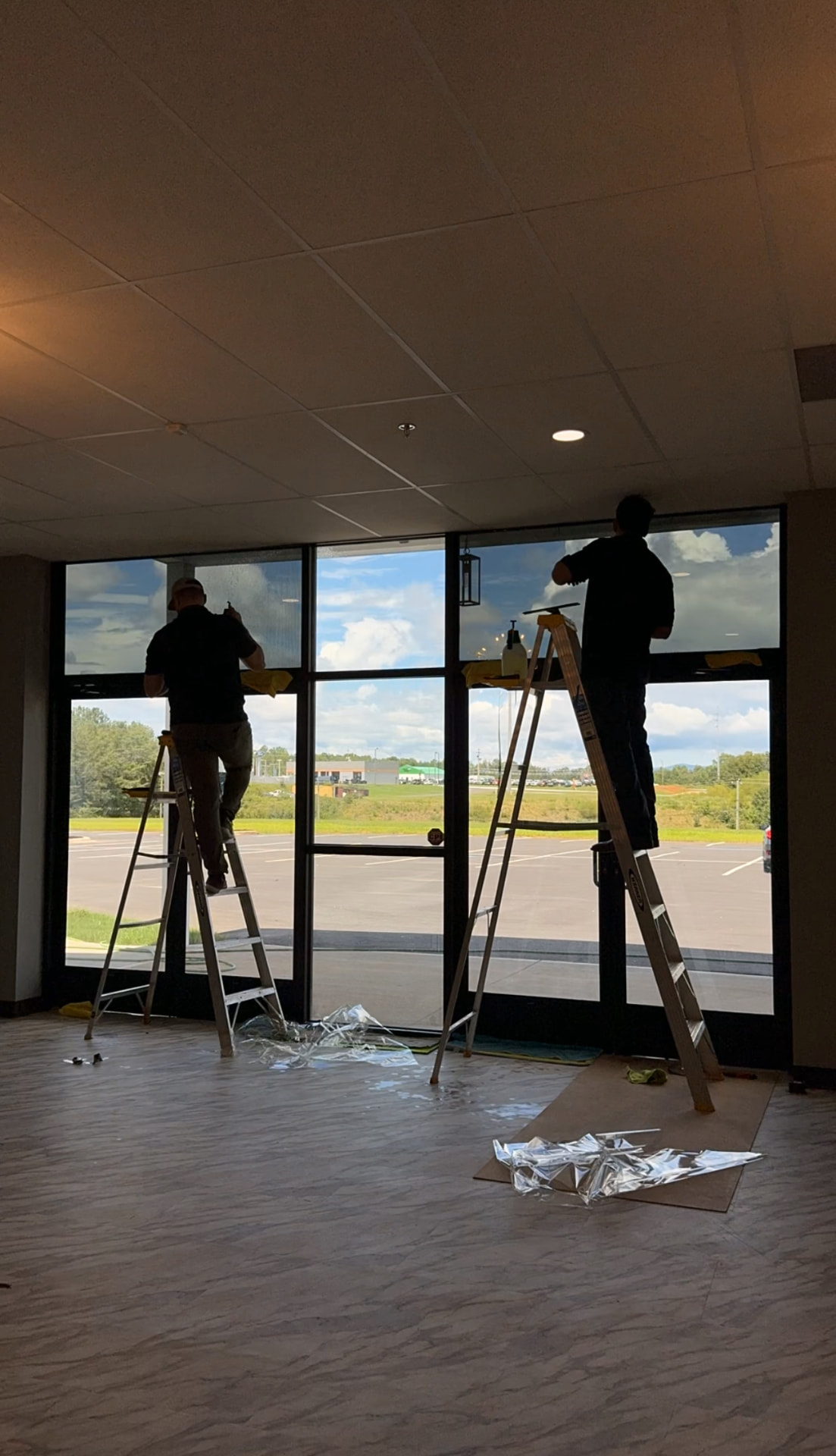 Two installers on ladders applying film to storefront glass inside an occupied building